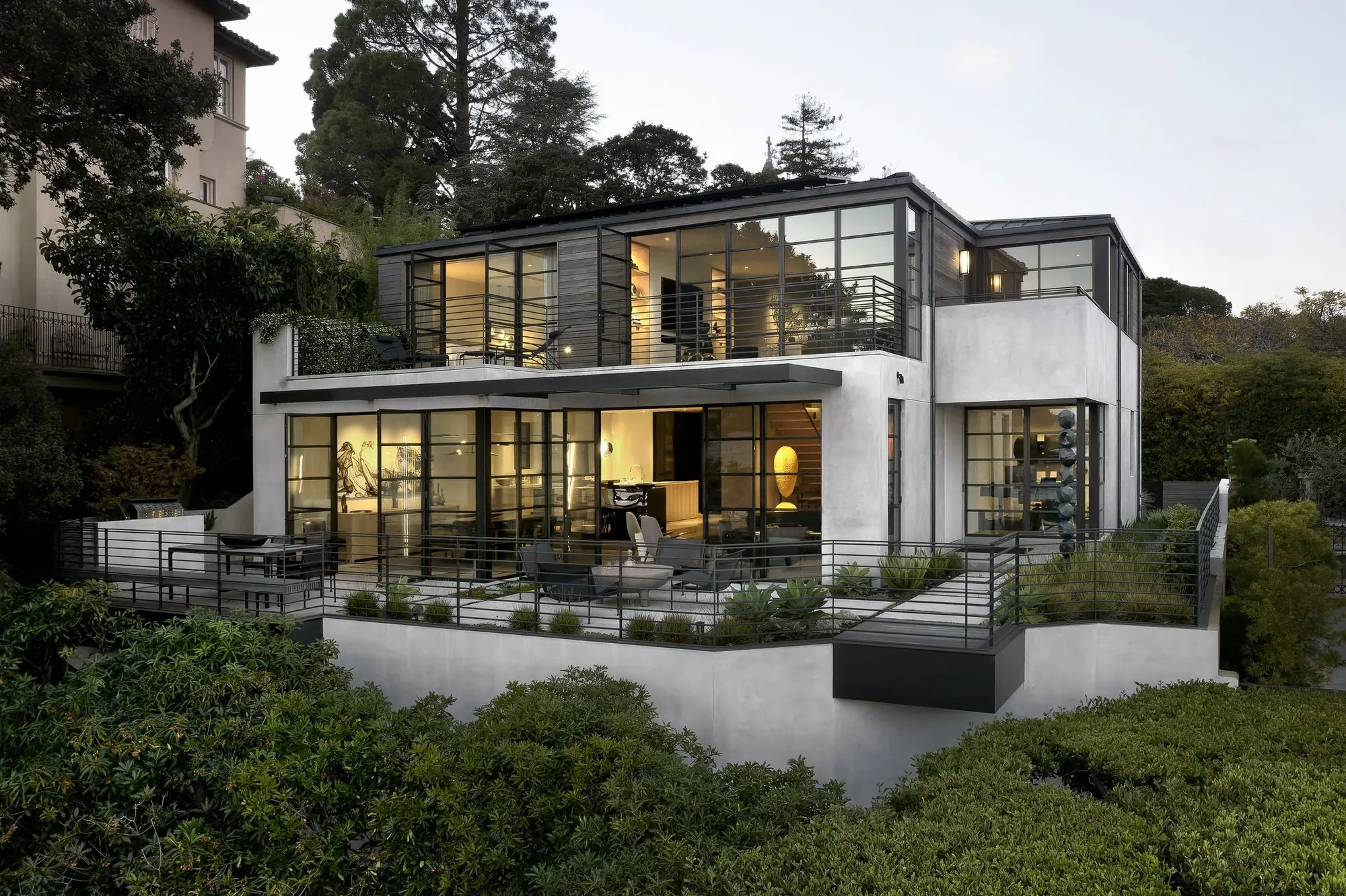 The house from below at dusk — lit glazing above a planted bank, the terrace running the length of the living room