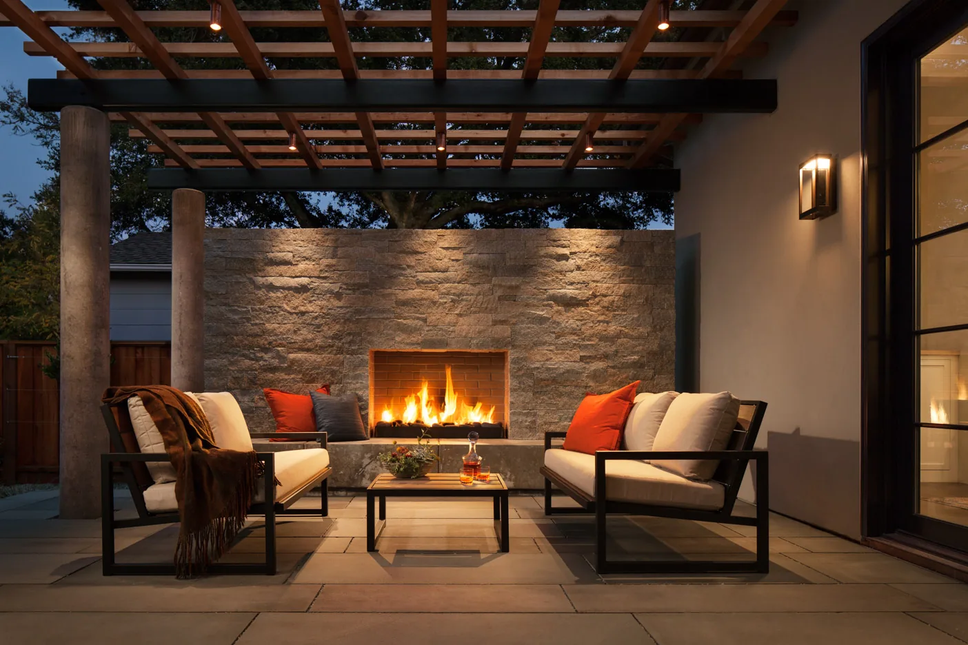Lafayette Residence — the outdoor living room at dusk, a lit stone fireplace under a timber pergola