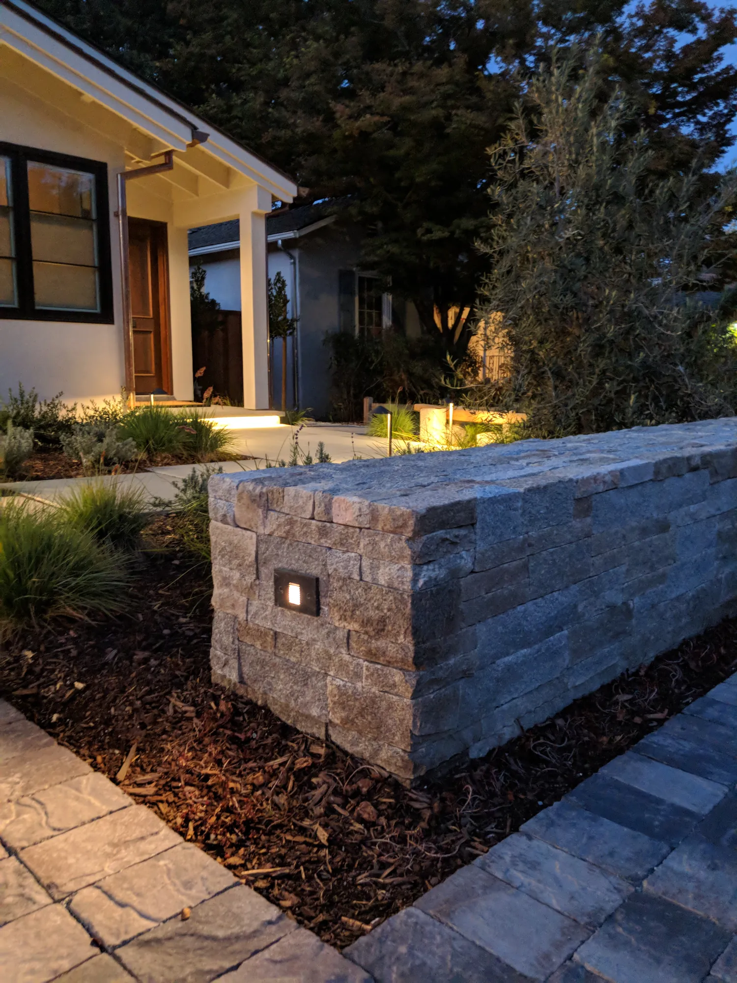 Lafayette Residence at dusk — the low dry-stack stone wall glowing at the arrival, with the lit entry beyond