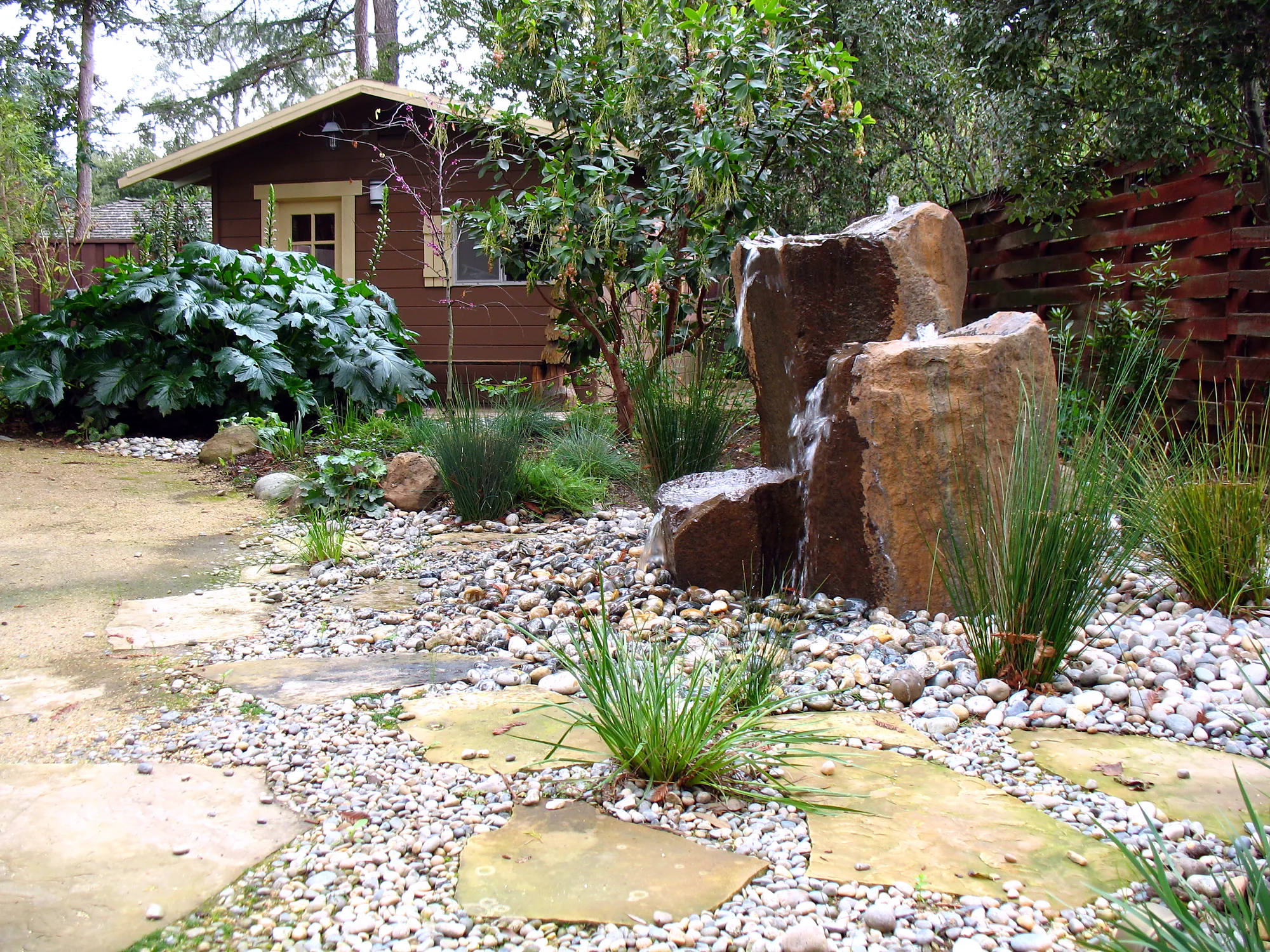 Los Altos family garden — the children's garden with a basalt-column fountain over river pebbles, rushes, and a small brown garden shed