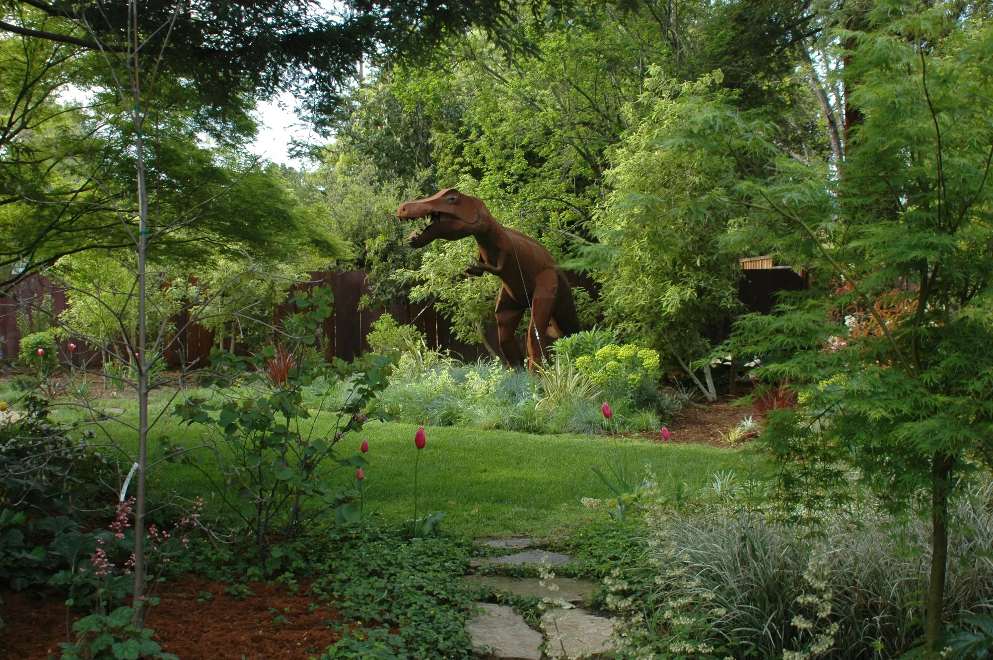 Los Altos Residence — the rusted-steel dinosaur standing among the planting above a small lawn, with a flagstone path and spring tulips