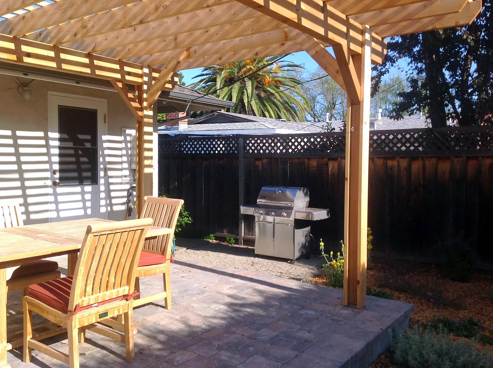 After — the same corner as a paver terrace under the new pergola, dining chairs in the dappled shade