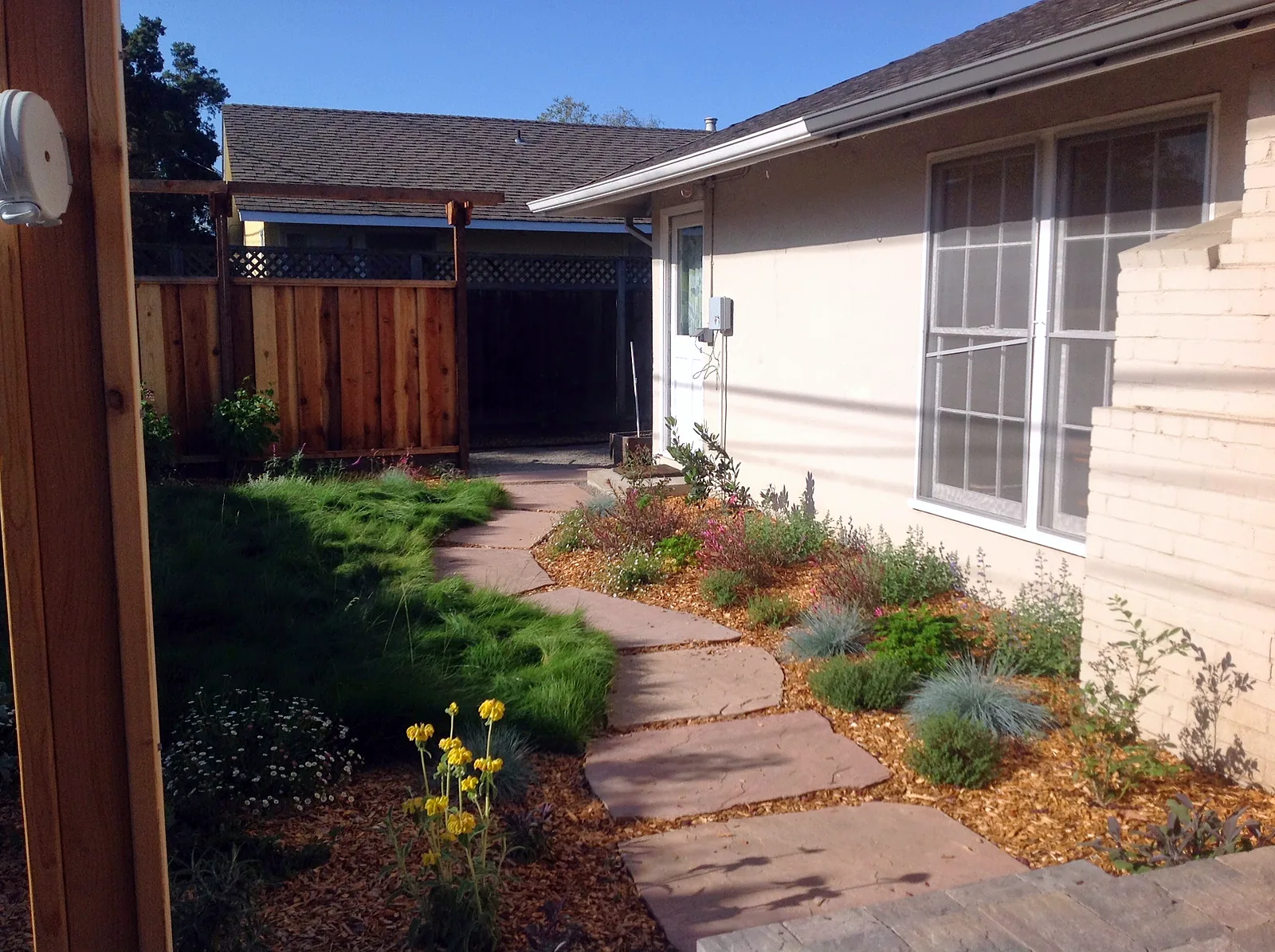 A flagstone path through meadow grass along the side of the house, with a mulched border of natives and herbs at the foundation