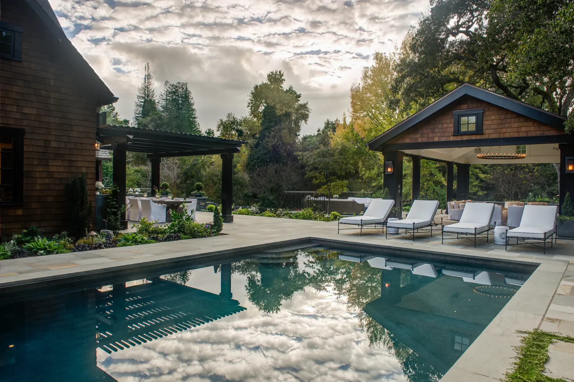 Orinda Residence — the rebuilt pool reflecting a cloud-streaked sky, with the open-air pavilion at its far end and a pergola beside the house