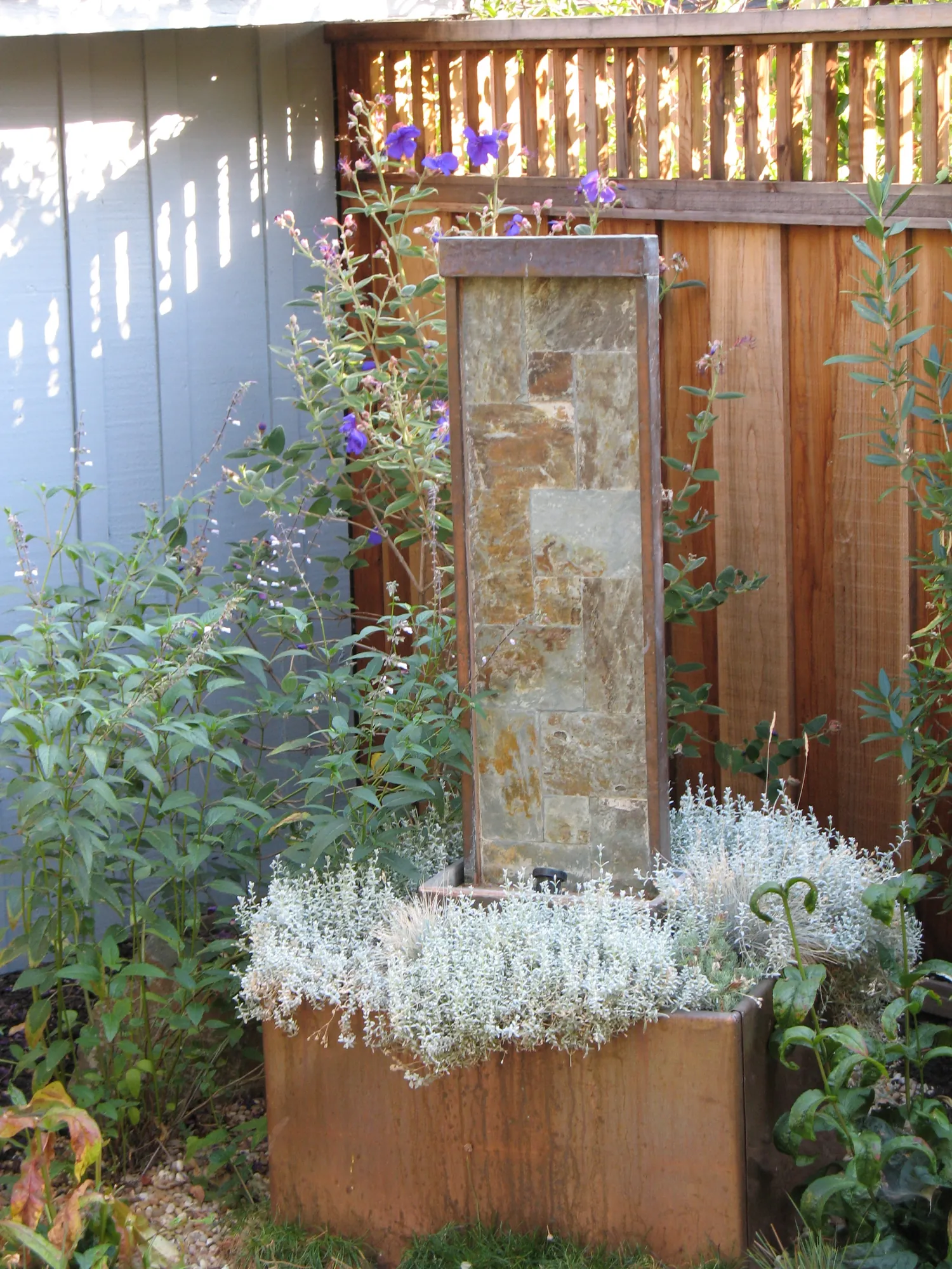 Slate-and-copper column fountain rising from a copper basin ringed with silver santolina, princess flower blooming behind