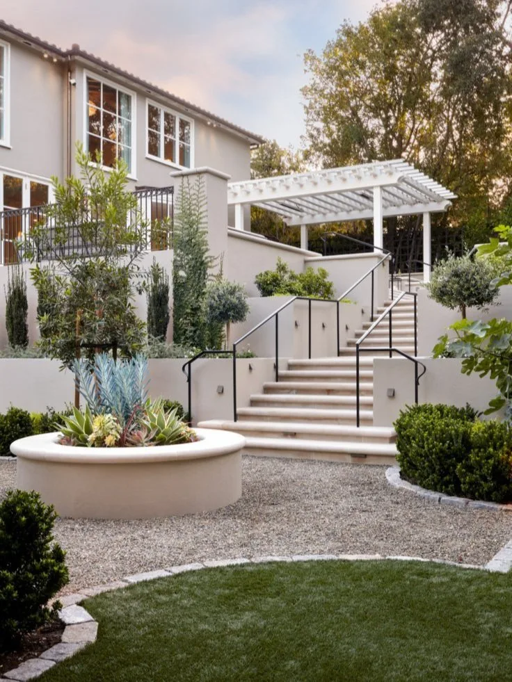 Dusk over the terraced garden — curved plaster stairs and black iron rails climbing to the white pergola and the lit house