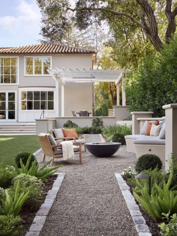 A cobble-edged gravel path between fern borders, leading past a dark fire bowl toward the lawn and pergola