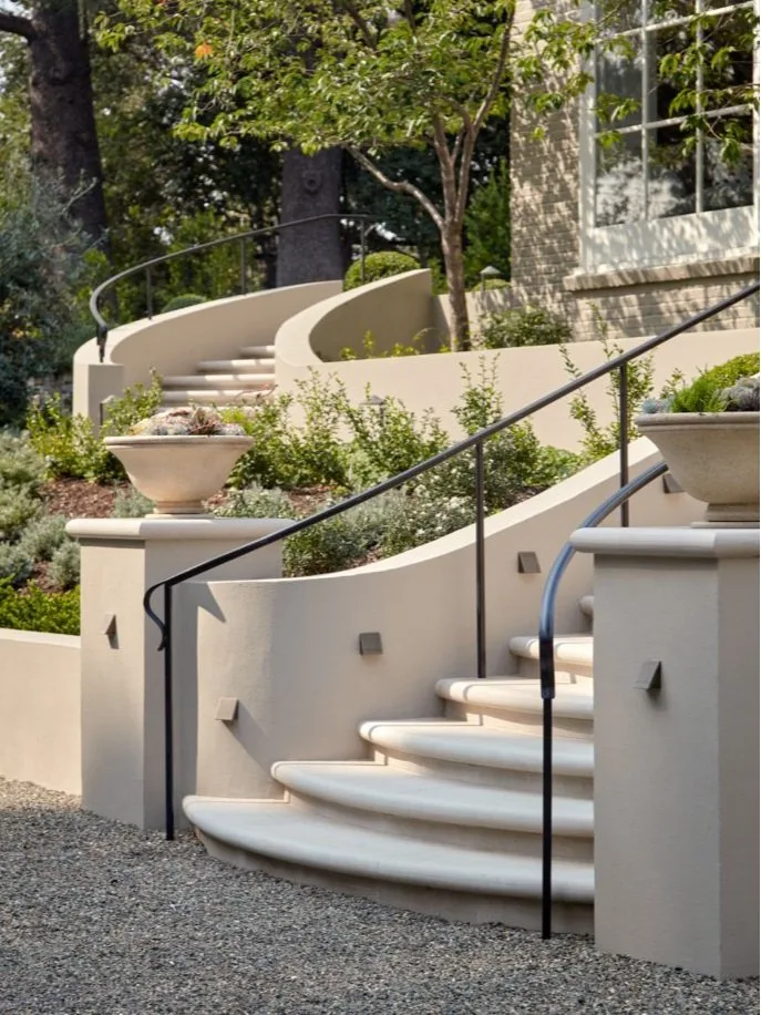 The curved entry stair — plaster walls with stone urns and a sweeping black iron handrail rising from the gravel court