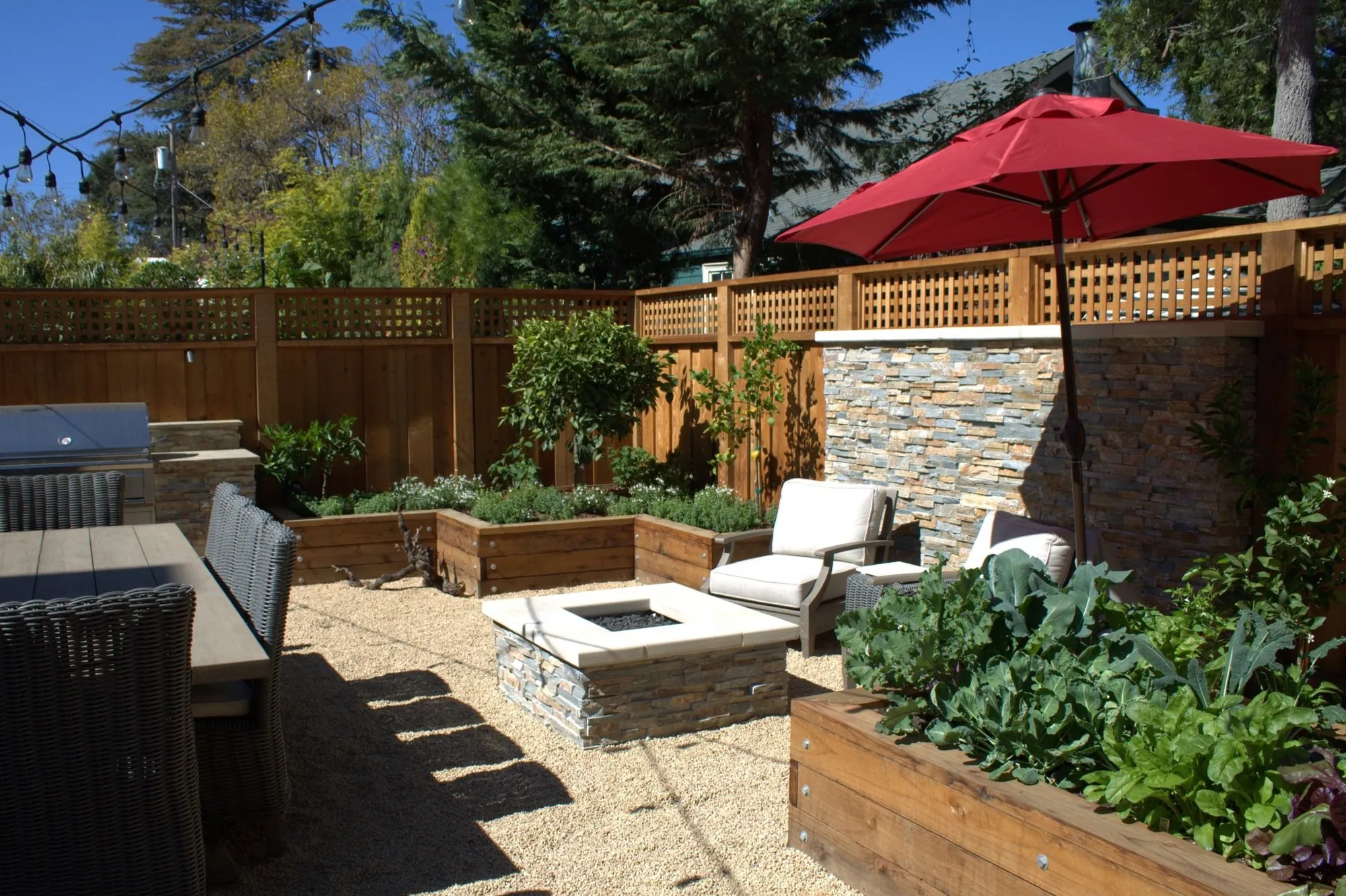 Burlingame Residence — a stacked-stone fire pit and lounge chairs on decomposed granite, ringed by raised vegetable beds under a red umbrella