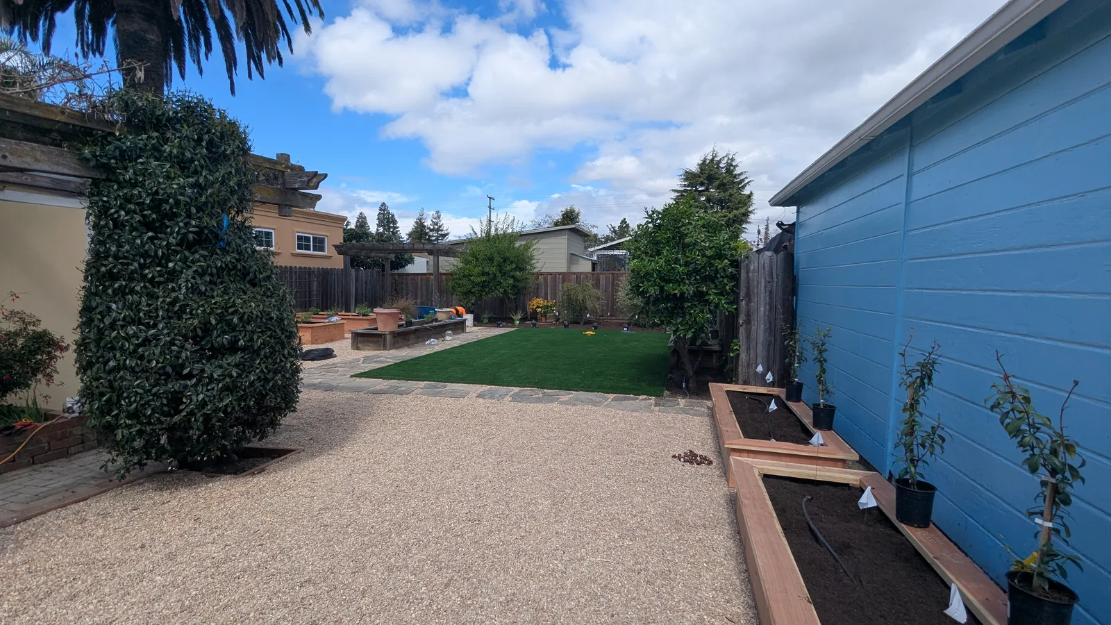 After photo — the same view, freshly built: a stabilized gravel terrace meeting a pet-friendly synthetic lawn on a flagstone border, with new raised planter beds along the garage