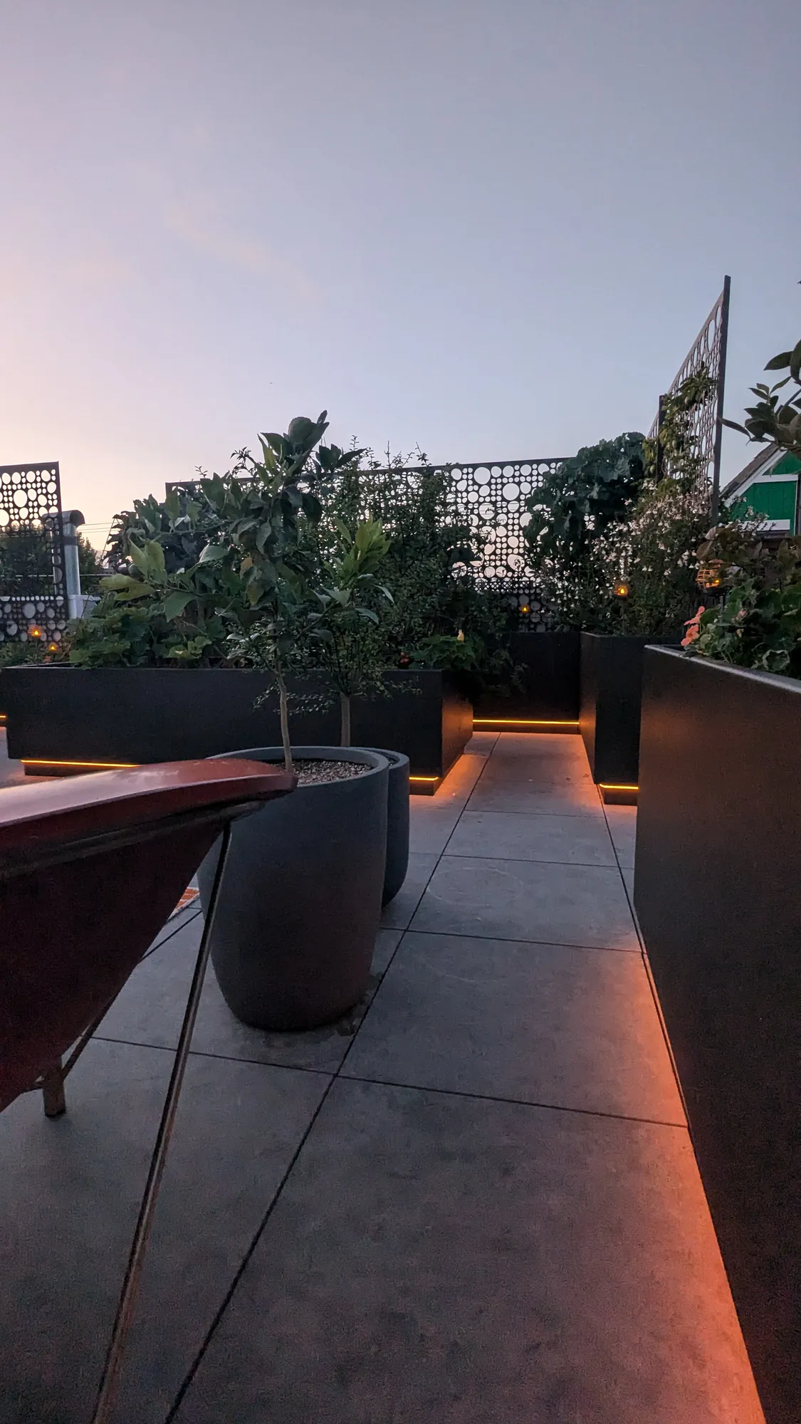 San Francisco rooftop edible garden at dusk — raised planters glowing with warm LED light beneath a pink sky, laser-cut metal privacy screens silhouetted against the city