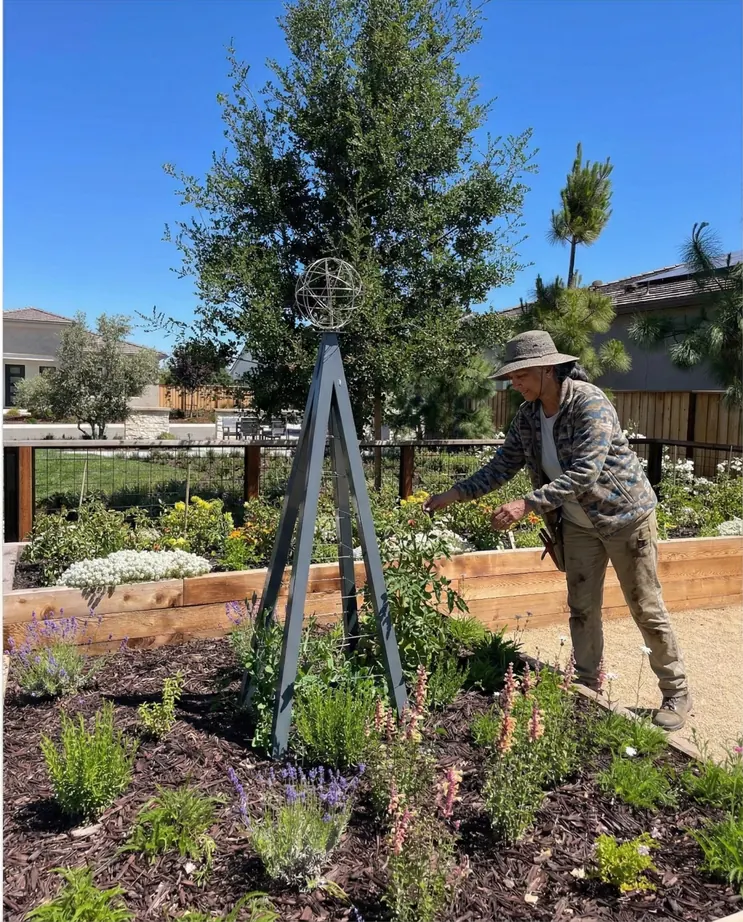 Tracy Lakes Agrihood — a gardener tending zinnias beneath one of the farm's signature steel trellises, with lavender, snapdragons, and pollinator planting in the surrounding beds
