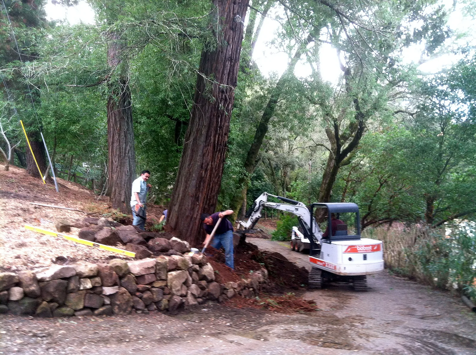 A crew building the wall along the drive, an excavator beyond